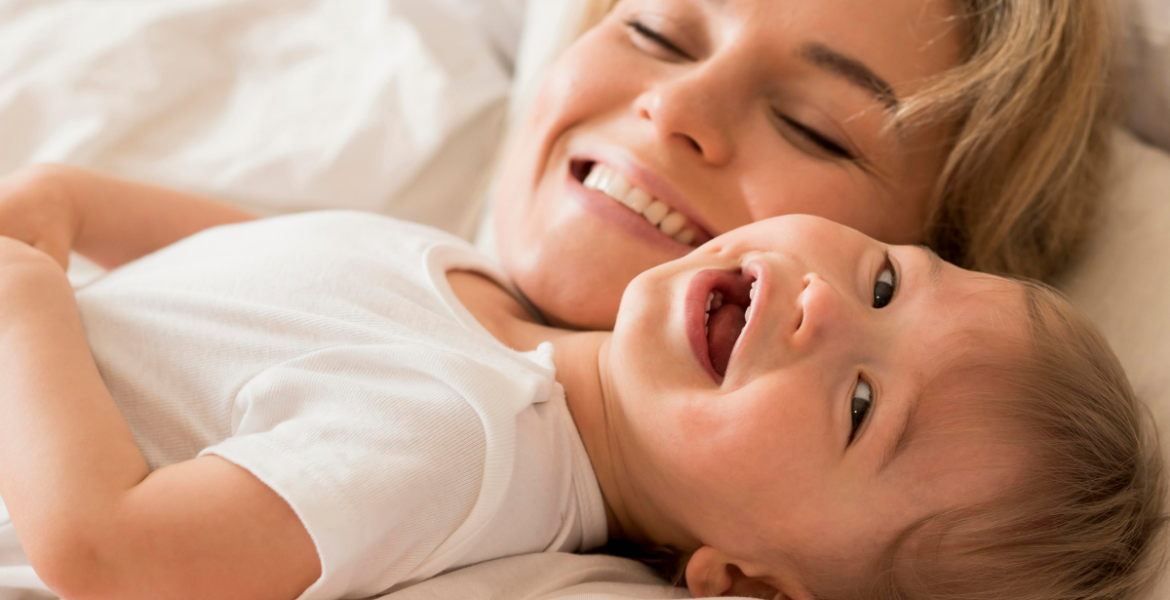 Madre sonriente abrazando a su bebé sobre una cama blanca, transmitiendo bienestar y ternura, ideal para comunicar el cuidado de la piel infantil