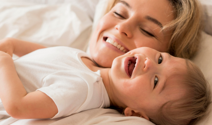 Madre sonriente abrazando a su bebé sobre una cama blanca, transmitiendo bienestar y ternura, ideal para comunicar el cuidado de la piel infantil