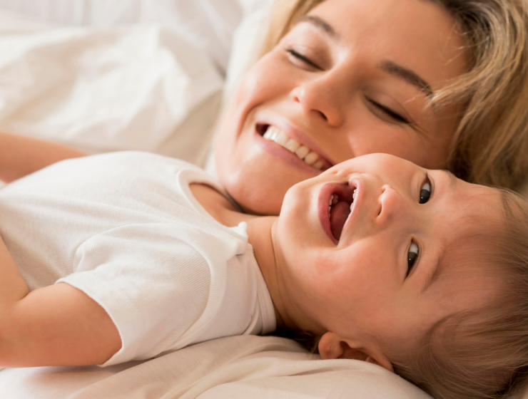 Madre sonriente abrazando a su bebé sobre una cama blanca, transmitiendo bienestar y ternura, ideal para comunicar el cuidado de la piel infantil