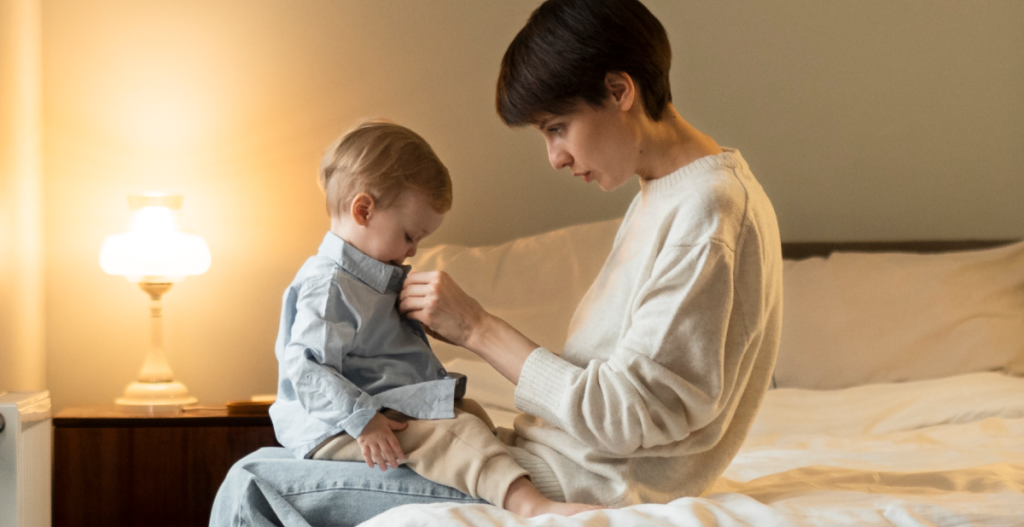 Madre sentada en una cama abrochando la camisa de su hijo pequeño con ternura, un momento cotidiano que refleja la rutina de cuidado de la piel infantil, ideal para niños con piel atópica que necesitan hidratación y protección suave
