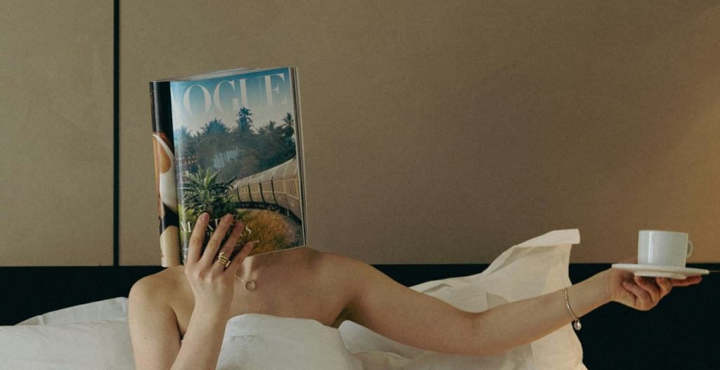 Persona tumbada en la cama, cubierta con una sábana blanca, sosteniendo una taza de café en una mano y una revista Vogue que le cubre la cara con la otra.
