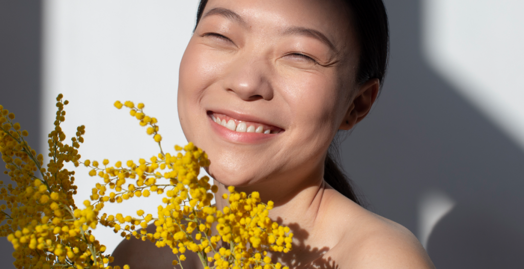 : Mujer sonriendo y sosteniendo flores amarillas bajo la luz natural, reflejando los beneficios de una piel cuidada