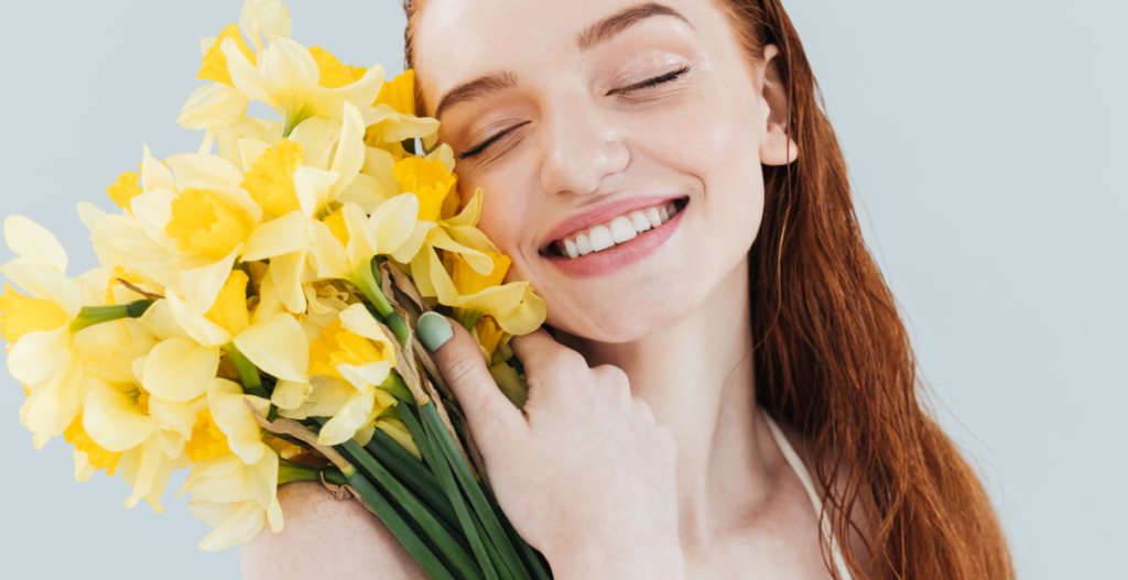 Mujer sonriente sosteniendo flores amarillas junto al rostro, evocando una rutina skincare por estaciones en primavera