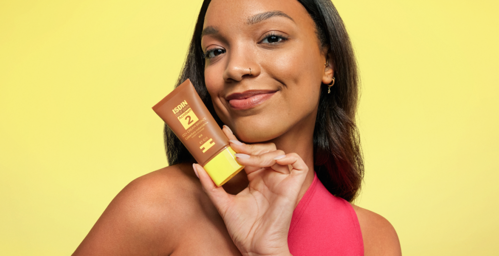 Mujer joven de piel morena sostiene Acniben CC+ Cream Bronze de ISDIN junto al rostro sobre fondo amarillo.