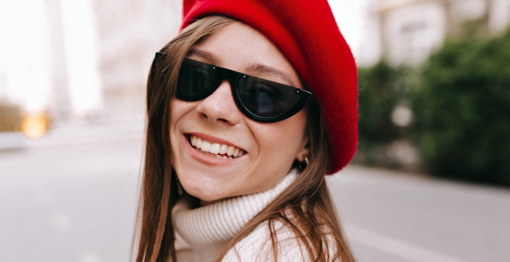Mujer joven sonriendo en exterior con gorro rojo y gafas de sol, transmitiendo estilo urbano y cuidado de la piel en invierno durante la Navidad