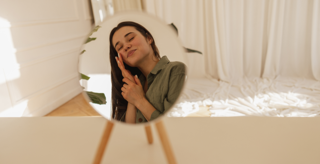 Mujer joven sonriendo con los ojos cerrados frente a un espejo circular, ambiente suave y luminoso. Concepto de autocuidado, skincare, propósitos y skin goals para 2026