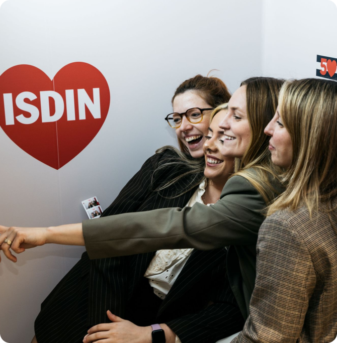 Cuatro amigas riendo mientras señalan un logo de corazón rojo en la pared. Momento espontáneo de amistad, metas 2026, skincare y celebración de skin goals compartidos.