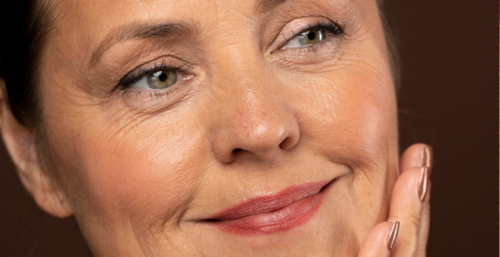 Mujer madura sonriendo con la mano en el rostro, mostrando arrugas de expresión como parte de una belleza natural y cuidada.