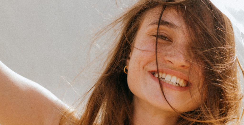 Mujer joven sonriendo al sol con el cabello suelto, mostrando una piel luminosa y natural.