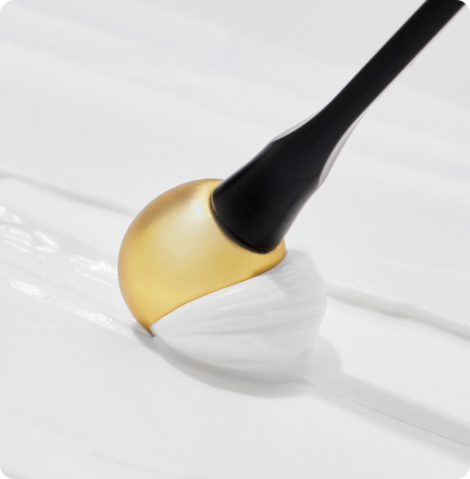 Aplicador dorado de contorno de ojos extendiendo una crema blanca con textura suave sobre una superficie lisa.