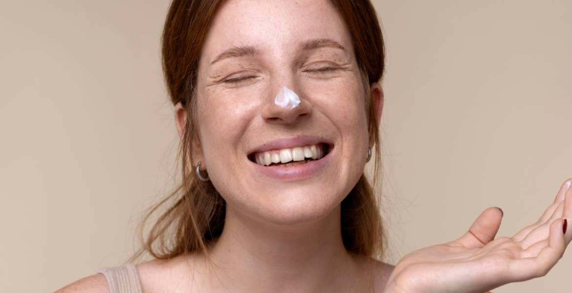Mujer con piel seca sonriendo y con crema hidratante en la nariz durante su rutina de cuidado facial.