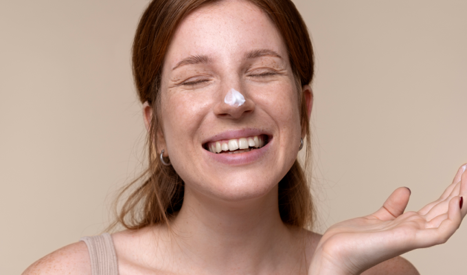 Mujer con piel seca sonriendo y con crema hidratante en la nariz durante su rutina de cuidado facial.