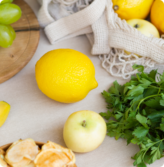 Limón, manzana y hierbas frescas sobre una superficie textil clara. Representa nutrición y hábito saludable para fortalecer uñas desde la alimentación, apoyando la estructura ungueal.