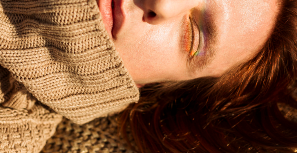 Primer plano de mujer pelirroja con suéter de lana beige, ojos cerrados y reflejo de arcoíris en el párpado, evocando calidez y luz otoñal.