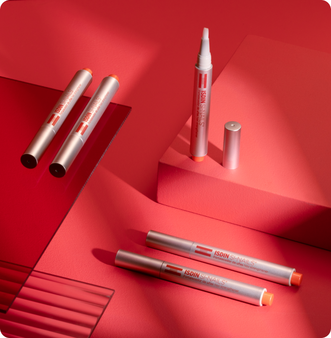Varios lápices fortalecedores de uñas ISDIN SI-Nails colocados sobre un fondo rojo con iluminación suave.