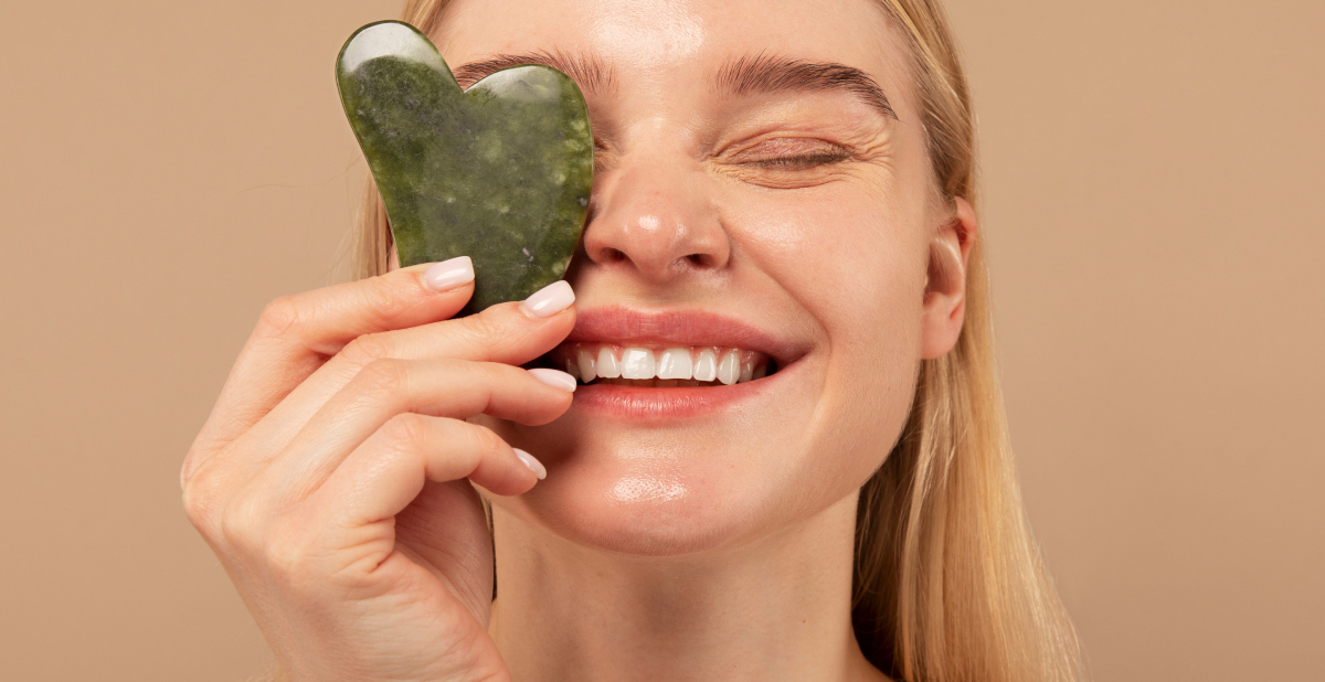 Mujer sonriendo mientras utiliza una piedra gua sha de jade verde para realizar un masaje facial.
