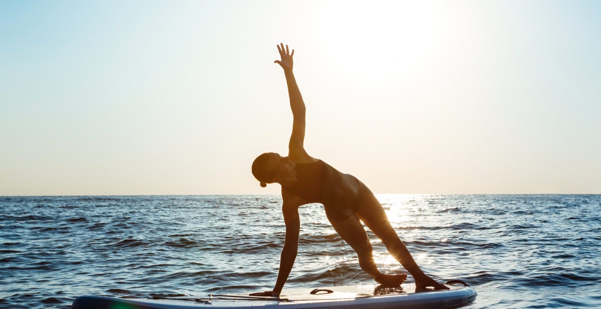 Silueta de una mujer practica yoga sobre una tabla de paddleboard en el mar al amanecer. Muestra equilibrio, mindfulness y ejercicio al aire libre como hábitos para la salud física y mental.