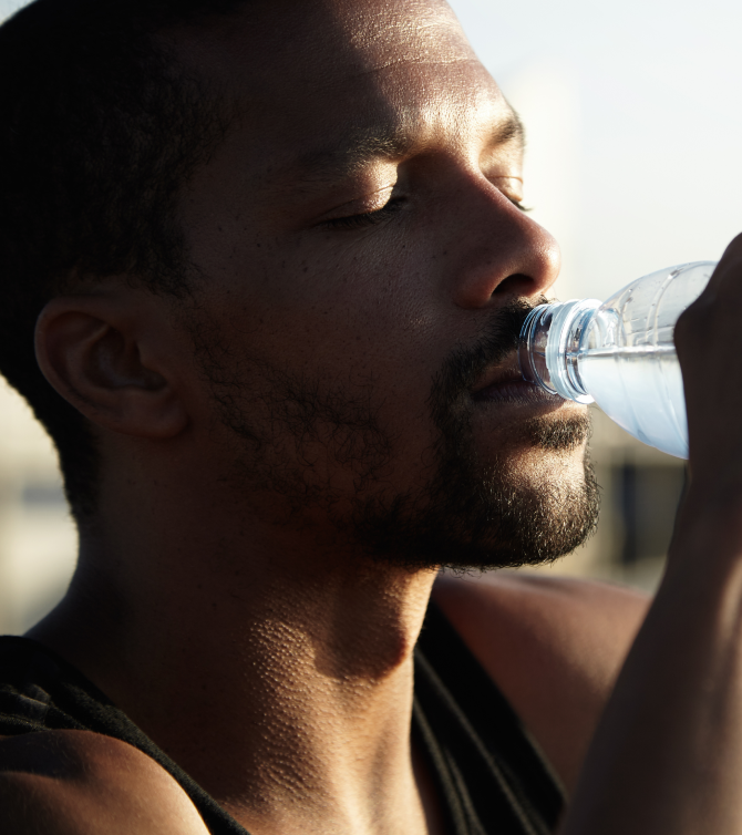 Hombre con barba bebe agua de una botella deportiva bajo luz cálida del sol. Representa hidratación, hábitos saludables y recuperación después del ejercicio.