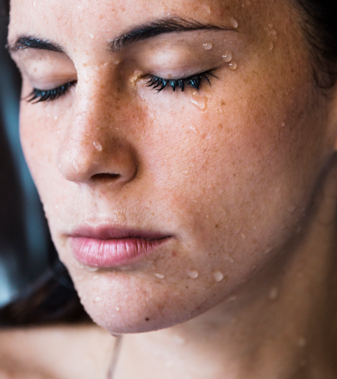 Primer plano de una mujer con los ojos cerrados y gotas de agua deslizándose por su piel. Evoca limpieza facial, autocuidado y rituales saludables como hábitos diarios de bienestar.
