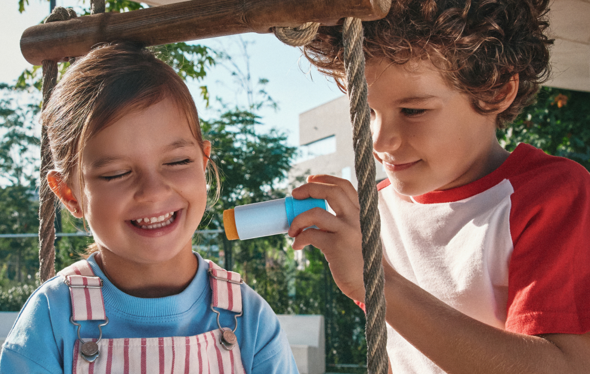 Dos niños juegan al aire libre mientras uno aplica un protector solar en stick a la otra.