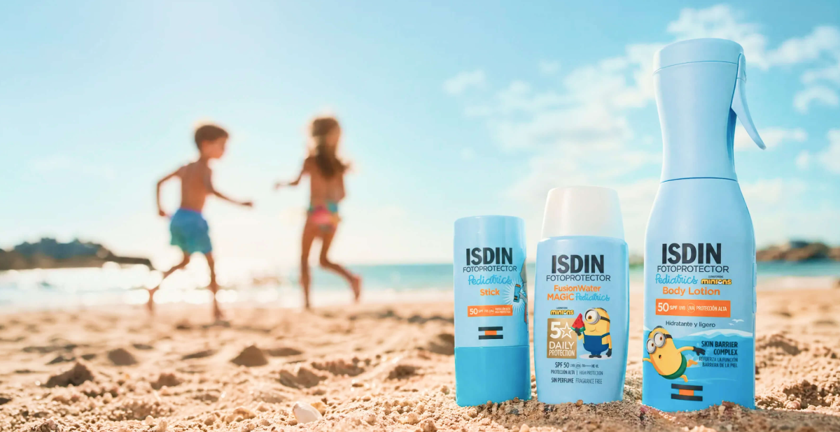 Tres fotoprotectores ISDIN Pediatrics con diseños de Minions colocados sobre la arena de la playa.