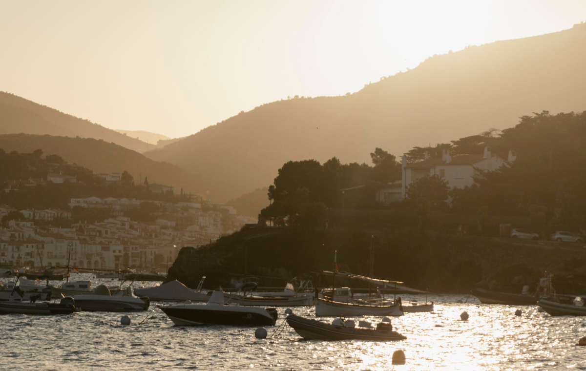 Bahía mediterránea con barcos fondeados y un pueblo costero entre montañas al atardecer