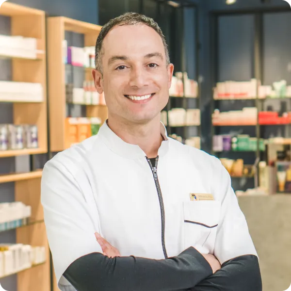 Eduardo Senante, farmacéutico apasionado por el cuidado de la piel, sonríe con los brazos cruzados dentro de Farmacia Senante, rodeado de estanterías con productos dermocosméticos.