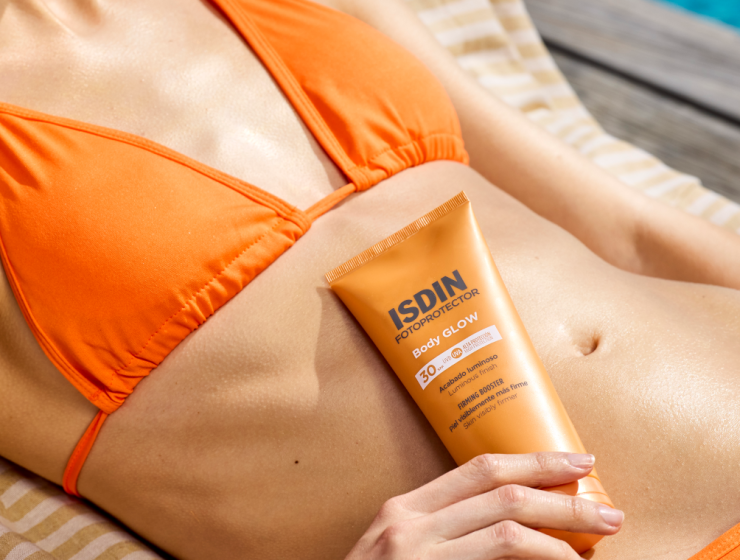 Primer plano de una mujer en bikini naranja recostada al borde de una piscina, sujetando ISDIN Body Glow SPF 30 sobre su abdomen luminoso y tonificado.