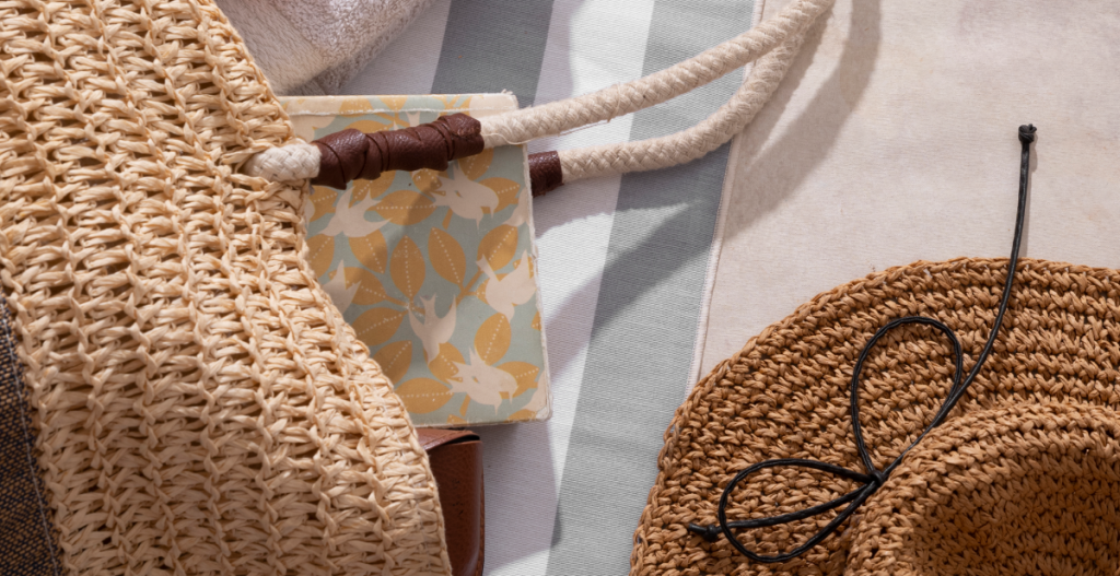 Imagen cenital de accesorios veraniegos: bolsas de playa tejidas, sombrero de paja con cordón negro, toalla y una libreta con estampado floral.