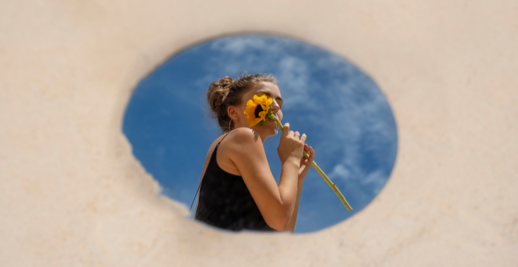 Toma artística de una mujer con un girasol reflejada en un espejo redondo colocado sobre arena, con el cielo azul de fondo.
