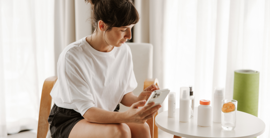 Mujer sentada en casa, observando su teléfono móvil frente a una mesa con productos cosméticos y un vaso de agua con rodajas de naranja.