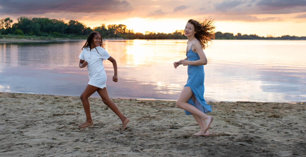 Dos mujeres corren descalzas por la arena junto al mar durante el atardecer, disfrutando del momento con luz dorada reflejada en el agua.