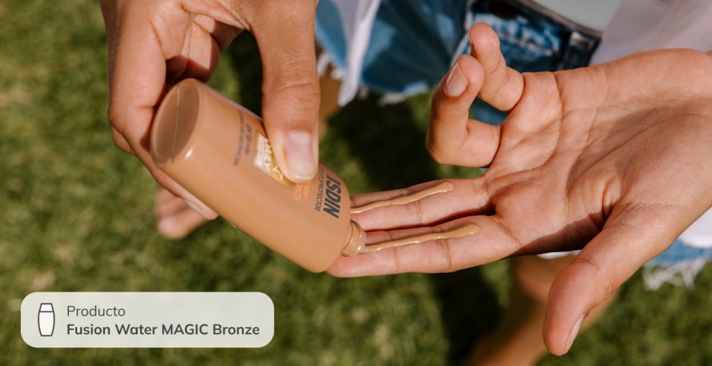 Primer plano de unas manos aplicando protector solar ISDIN Fusion Water MAGIC Bronze al aire libre, sobre fondo de césped.