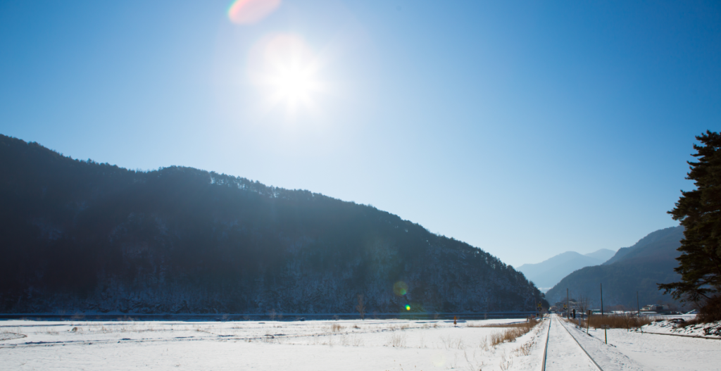 Montañas y nieve bajo sol fuerte, vía de tren hacia el horizonte. Concepto: cuidado de la piel en invierno, mitos del sol frío vs. SPF todo el año, vibra de renovación de Año Nuevo.