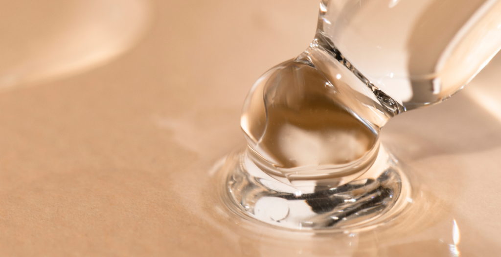 Gota de sérum transparente extendiéndose sobre una superficie, simbolizando activos hidratantes que apoyan la renovación celular de la piel.
