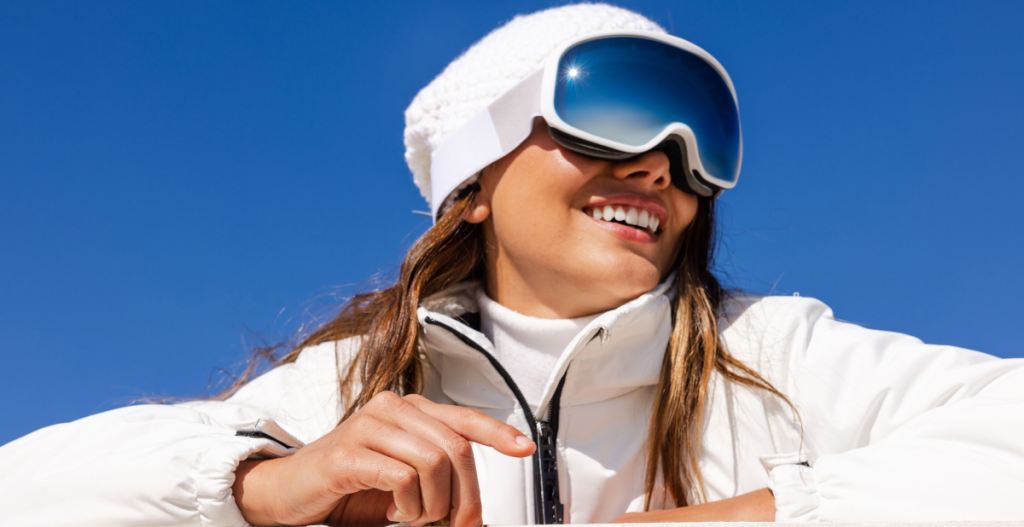 chica esquiando con gafas de sol que reflejan, fondo azul intenso y ropa blanca, está feliz y sonrie