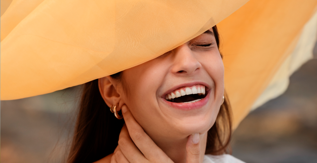 Mujer sonriendo con los ojos cerrados bajo una tela dorada iluminada por el sol, representando una piel luminosa y saludable