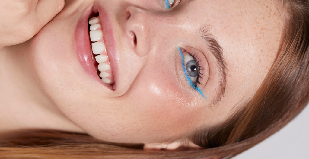 Mujer con piel natural y pecas llevando delineado azul sutil, ejemplo de maquillaje creativo con efecto piel real dentro de la tendencia make up no make up.