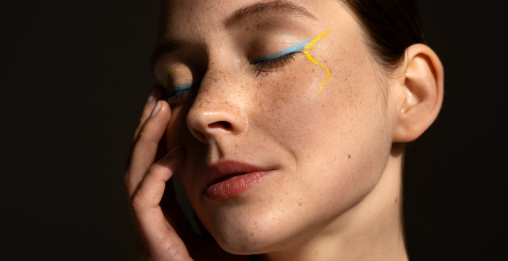 Primer plano de mujer con piel luminosa, pecas visibles y delineado azul suave que resalta la mirada en un maquillaje natural de efecto buena cara.