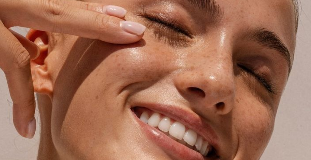 Persona sonriendo mientras aplica crema cerca del contorno de ojos, con la piel luminosa y cuidada.