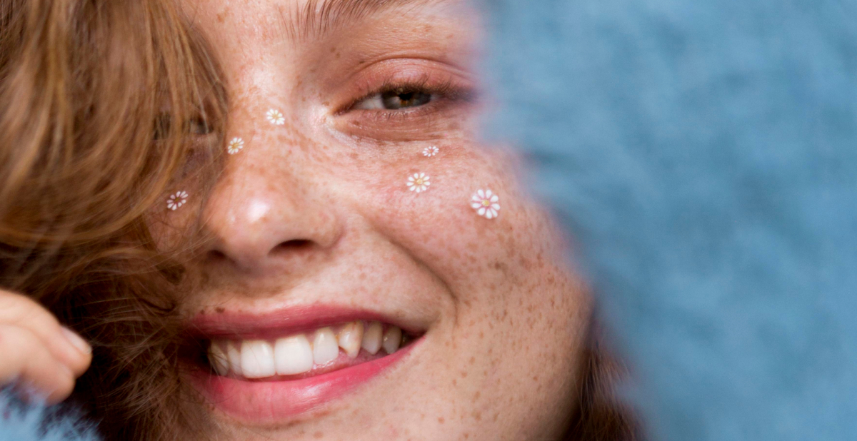 Persona sonriente con pecas y pequeñas flores blancas decorativas sobre el rostro.