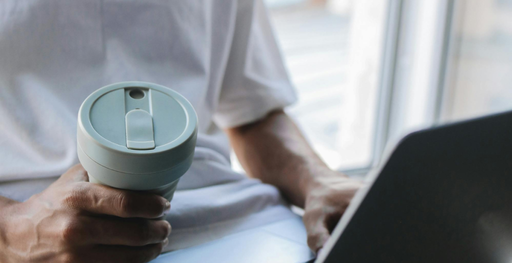 A person holding a reusable cup of coffee while working on a laptop near a window.