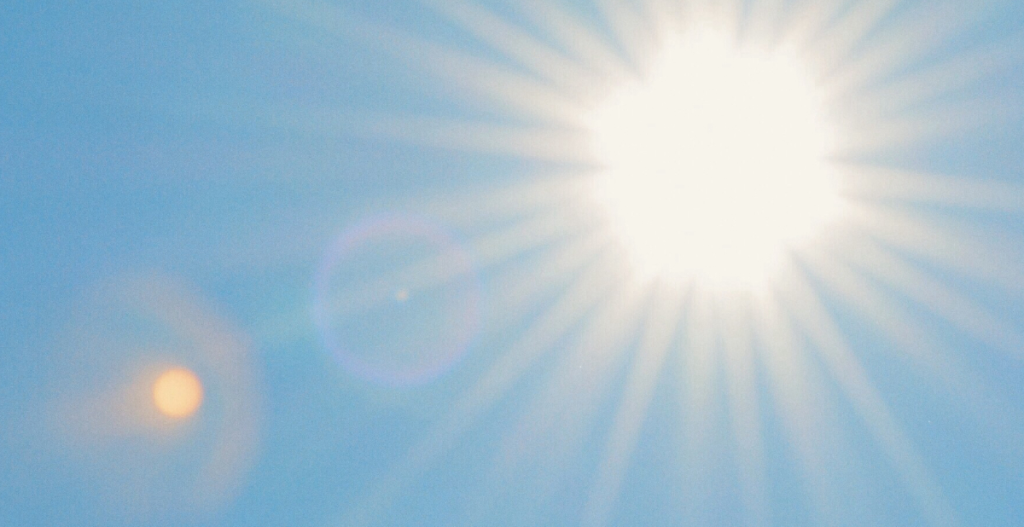Bright sun shining in a pure blue sky, with flames of lenses and visible rays.