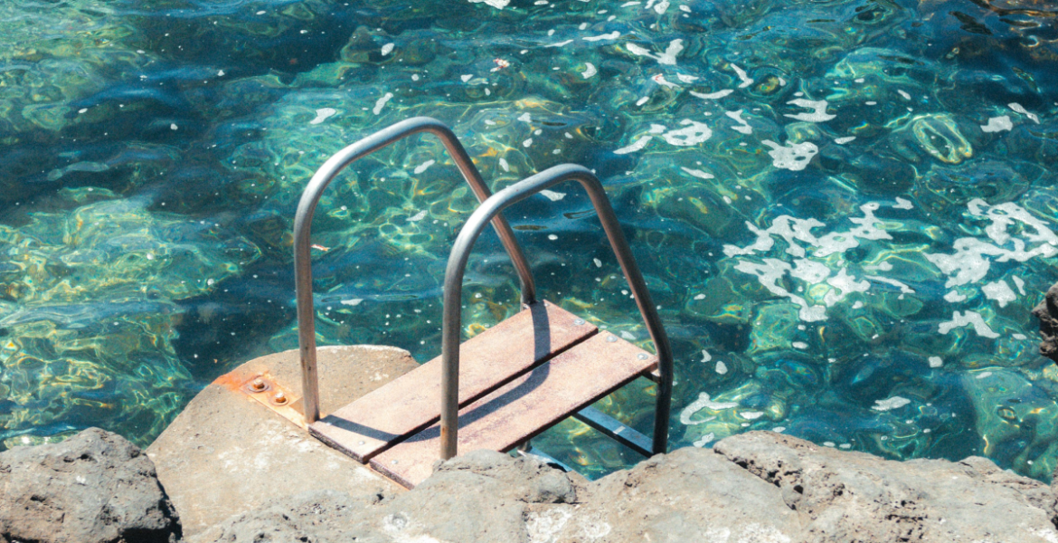 Metal ladder into mediterranean sea — A weathered metal ladder leading into crystal-clear turquoise water surrounded by volcanic rock.