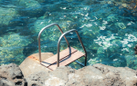 Metal ladder into mediterranean sea — A weathered metal ladder leading into crystal-clear turquoise water surrounded by volcanic rock.