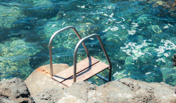 Metal ladder into mediterranean sea — A weathered metal ladder leading into crystal-clear turquoise water surrounded by volcanic rock.